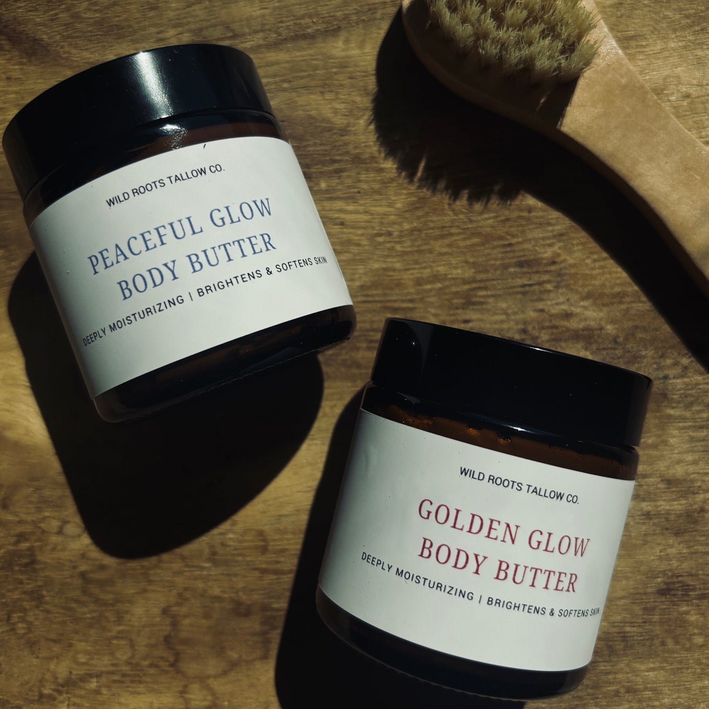Two jars of body butter labeled 'Peaceful Glow' and 'Golden Glow' on a wooden surface with a brush.